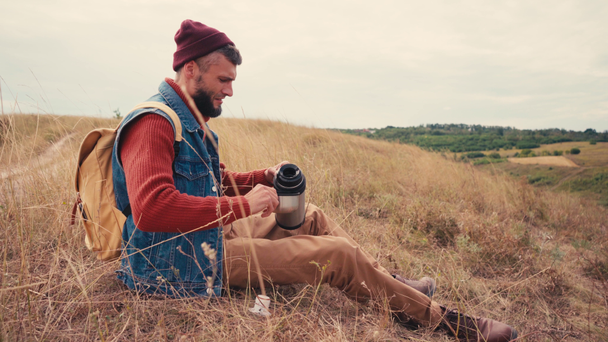 Reisender setzt sich hin und gießt Tee aus Thermoskanne auf Hügel - Filmmaterial, Video
