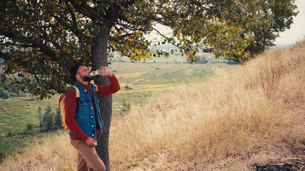 Podróżnik wspinający się na wzgórze, opierający się o drzewo i wodę pitną - Materiał filmowy, wideo