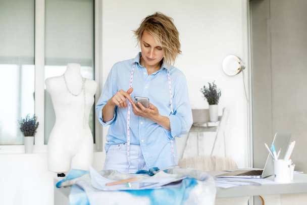 Mulher empresária no salão de vestir usando telefone e laptop, se comunica com o cliente, ferramentas de tricô e esboços na mesa. - Foto, Imagem