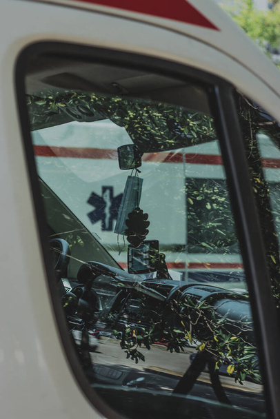 Вертикальный снимок салона скорой помощи в Сплите, Хорватия. Припаркован на общественной парковке, отражение деревьев на окне. Лицевая маска, свисающая с зеркала заднего вида - Фото, изображение