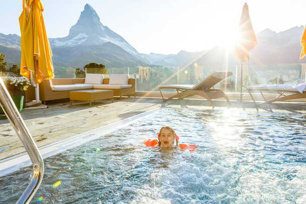 アルプスの山々、オーストリアの豪華なスパアルプスリゾートの屋外スイミングプールで遊んでいる小さな女の子。冬と子供と雪の休暇。山の景色を望むホットタブ屋外。子供たちが遊んだり泳いだり. - 写真・画像