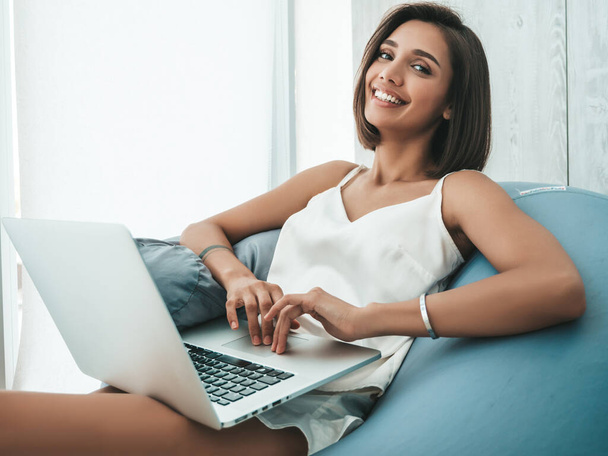 Portrait of beautiful smiling woman dressed in white pajamas. Carefree model sitting on soft bag chair and using laptop. Sexy female reading news and enjoying her morning at balcony - Foto, immagini
