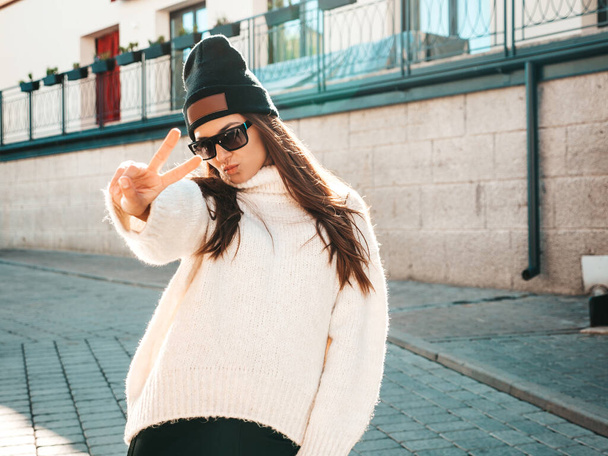 Portrait of beautiful smiling model. Woman dressed in warm hipster white sweater and beanie. She posing in the street at sunset in sunglasses - Foto, afbeelding