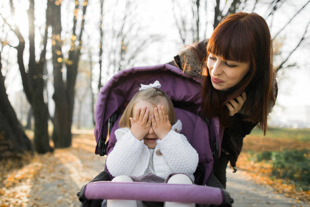 Walk in autumn park. Happy family, young mother walking with her little cute baby girl and carries it in a pink pram. Pretty baby girl hiding her eyes and face with hands - 写真・画像