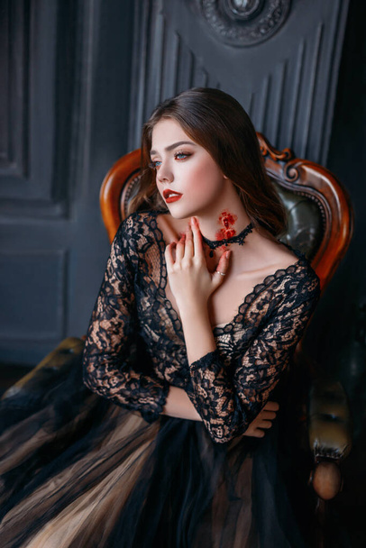 beautiful adult girl, victim of a vampire with a bloody bite on her neck. Young woman in a black gothic dress. Creative makeup and carnival costume for a halloween party. Dark room background - Foto, Imagem