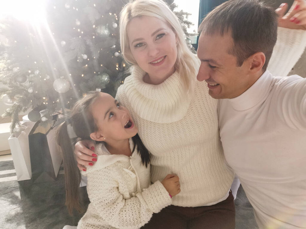 семья, праздники, технологии и люди - улыбающиеся мама, папа и маленькая девочка, делающие селфи с фотоаппаратом над гостиной и фоном новогодней елки - Фото, изображение