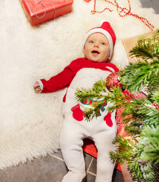 Cute baby little toddler boy lay on the carpet with open mouth laughing and looking at camera under Christmas tree - Foto, Imagem