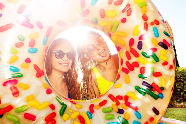 Two teenagers girls stick out tongues posing inside inflatable doughnut wearing sunglasses and smile - Foto, afbeelding