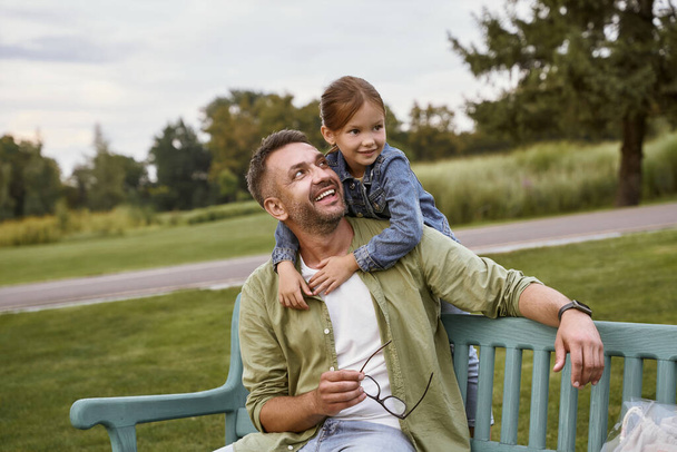 屋外で一緒に時間を過ごす。公園の木製のベンチに座って幸せな愛する父とかわいい娘の肖像画、抱擁と笑顔 - 写真・画像