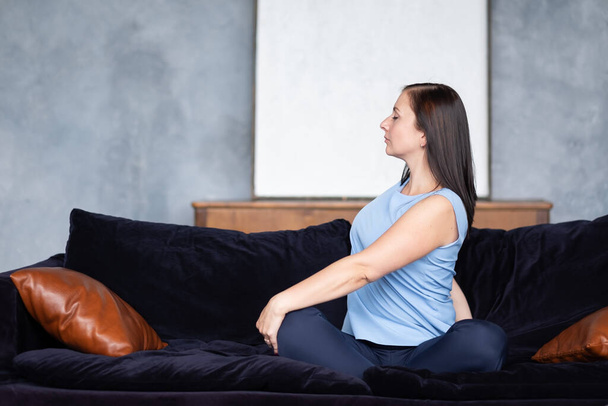 Młoda kobieta robi kręgosłup kręgosłupa pozy siedzi w domu na kanapie. - Zdjęcie, obraz