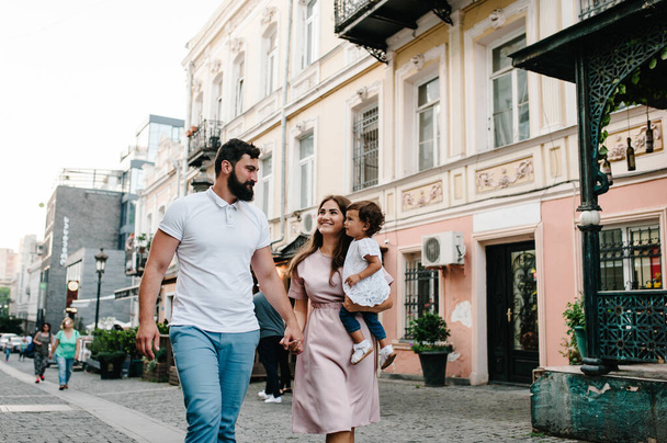 Young happy family: father, mother and daughter during a walk the streets of the old city, town, outdoors. The concept of family holiday and travel. The mom, dad holds and hugs little girl. - Fotoğraf, Görsel