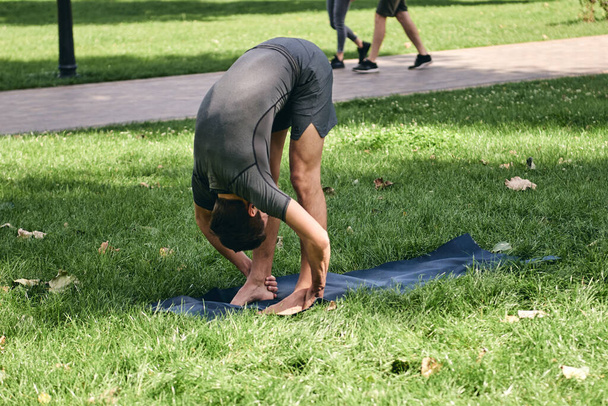 Urheilupukuinen nuori mies joogaa puistossa. Harjoitus seisoo eteenpäin mutka Asana aiheuttaa ulkona. Ihmiset treenaavat vihreällä ruoholla joogamatolla. Käsite rauhallinen ja meditaatio. - Valokuva, kuva