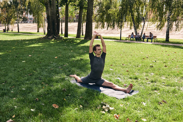 Young athletic man in sportswear doing yoga in the park. Practice asana outdoors. People exercising on green grass with yoga mat. Strong mature caucasian man in split pose. - 写真・画像