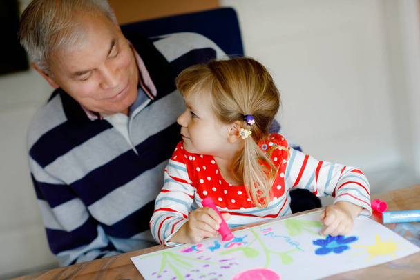 Cute little baby maluch dziewczyna i przystojny starszy dziadek malarstwo z kolorowe filcowe długopisy i ołówki w domu. wnuk i mężczyzna dobrze się razem bawią, kreatywna rodzina. - Zdjęcie, obraz
