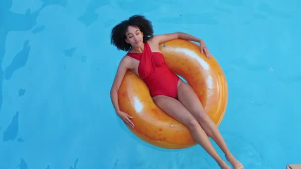 Tanned curly teenage girl in fashionable swimsuit relaxing in pool after school on vacation at home in the spa, touching cool wet water with hands, reclined in orange swimming lifebuoy ring, top view - Footage, Video