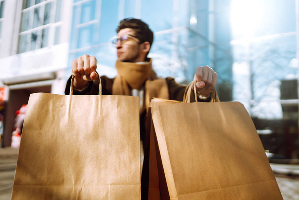 Hand of young man with paper bags with purchases. Stylish man holds out shopping bags on the city street. Consumerism, purchases, shopping, black friday, lifestyle concept. - Фото, изображение