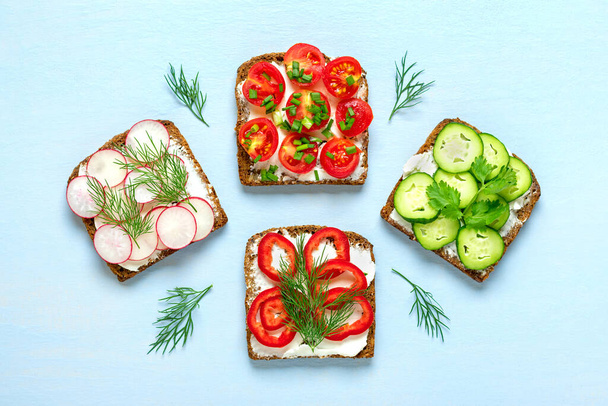 Variety of sandwiches for breakfast - slice of whole grain dark bread, red pepper, cream cheese, cucumbers, radishes, cherry tomatoes, garnished with dill, green onions on blue table Top view Flat lay. - Photo, Image