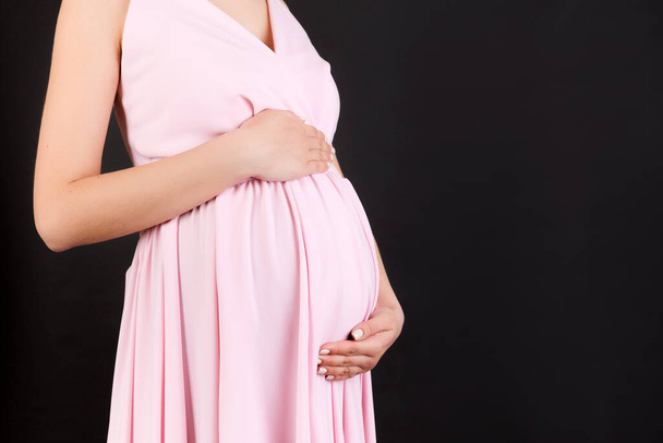 Zugeschnittenes Bild einer schwangeren Frau in rosa Kleid. Die junge Mutter umarmt ihren Bauch und erwartet vor schwarzem Hintergrund ein Baby. Kopierraum. - Foto, Bild