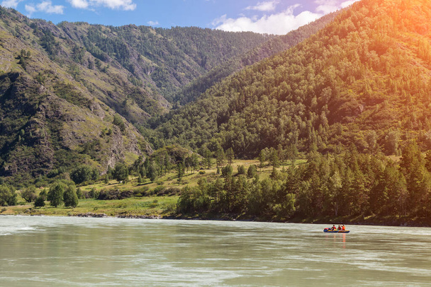 Turistická zábava v Altai - rafting po řece na nafukovací lodi na pozadí hor pokryté zelenou trávou a stromy a modrá obloha. - Fotografie, Obrázek