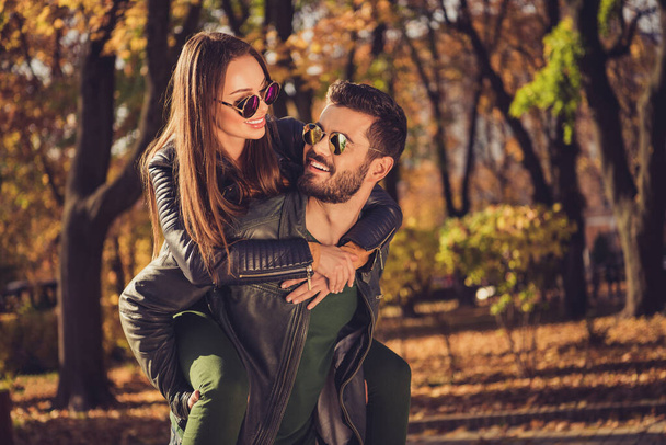 Photo of positive harmony couple guy hug look piggyback charming girl in fall city foliage park wear sunglass jacket - Foto, Imagen