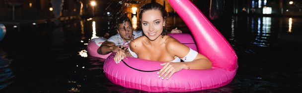 Sito web intestazione della giovane donna guardando la fotocamera mentre nuota con anello gonfiabile vicino al fidanzato in piscina di notte - Foto, immagini