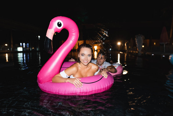 Szelektív fókusz a fiatal nő nézi kamera, miközben úszás gyűrű közelében barátja medence éjjel  - Fotó, kép