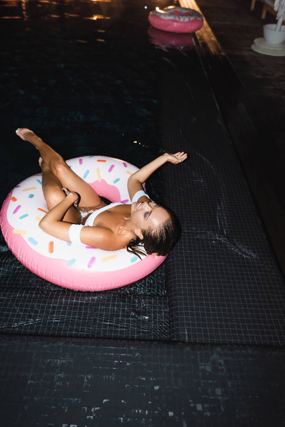 Selective focus of young woman lying on swim ring in pool at night  - Foto, Imagen