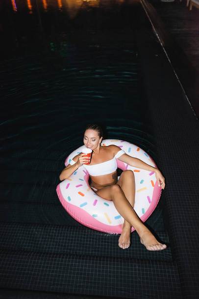 Mladá žena drží jednorázové pohár, zatímco sedí na plaveckém kroužku v bazénu v noci  - Fotografie, Obrázek