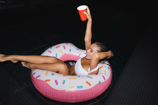 Brunette femme tenant tasse jetable tout en nageant dans l'anneau dans la piscine la nuit  - Photo, image