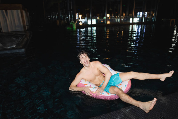 Excited muscular man sitting on swim ring in pool at night  - Foto, afbeelding