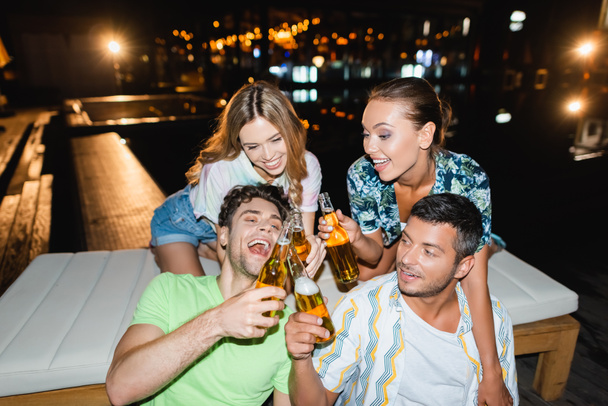 Young friends toasting with beer bottles near swimming pool at night  - Foto, Bild