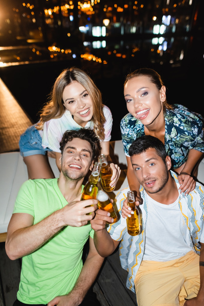 Young couples looking at camera while clinking beer near swimming pool at night  - Foto, Bild