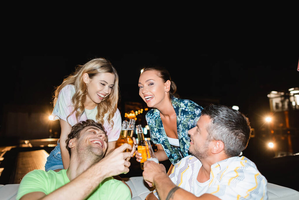 Selective focus of friends toasting with bottles of beer during party outdoors at night  - Fotografie, Obrázek