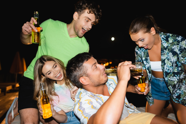 Mężczyzna pije piwo w pobliżu podekscytowanej dziewczyny i przyjaciół na zewnątrz w nocy  - Zdjęcie, obraz