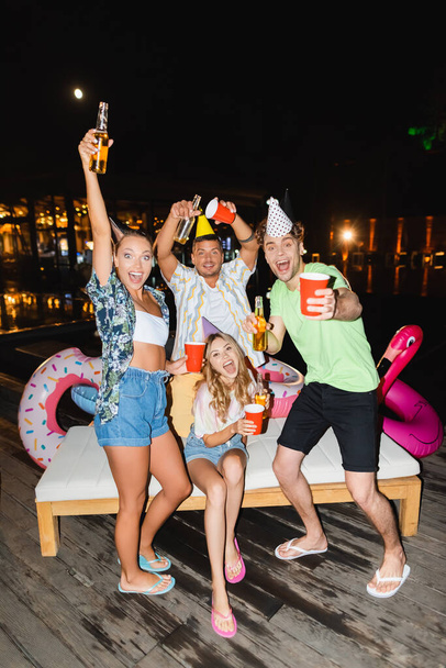 Jeunes amis en casquettes de fête tenant des bouteilles de bière et des tasses jetables à l'extérieur la nuit  - Photo, image