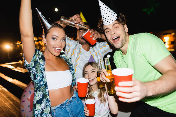 Concentration sélective des jeunes dans les casquettes de fête tenant des bouteilles de bière et des tasses jetables à l'extérieur la nuit  - Photo, image