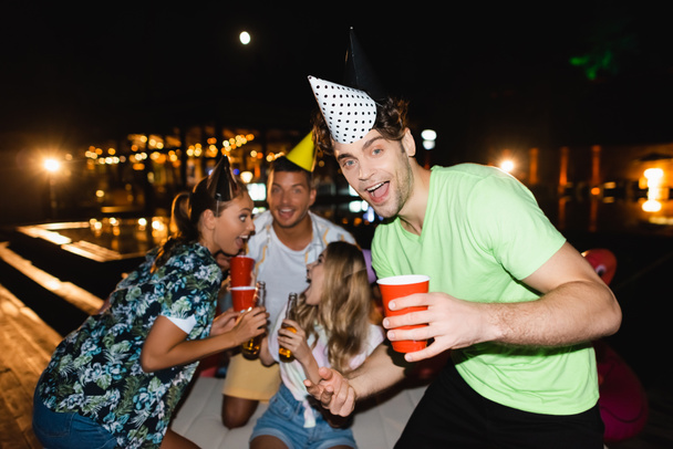 Selektywny skupienie młody człowiek w party cap trzymając jednorazowy kubek w pobliżu przyjaciół podczas imprezy w nocy  - Zdjęcie, obraz