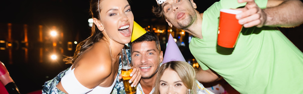 Foto panorámica de los jóvenes en gorras de fiesta mirando a la cámara durante la fiesta con cerveza por la noche  - Foto, Imagen