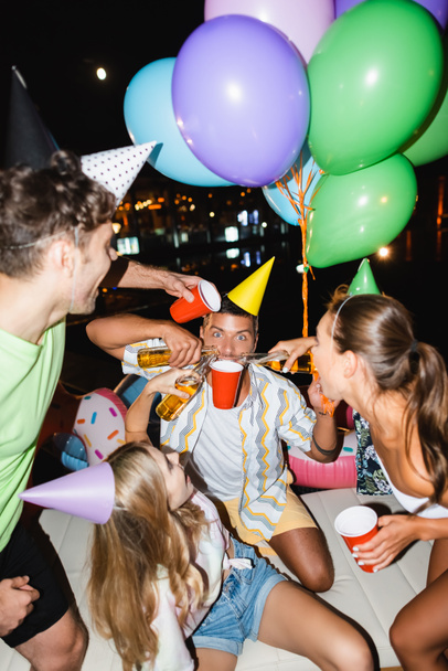 Enfoque selectivo del hombre sosteniendo taza desechable cerca de amigos vertiendo cerveza y globos al aire libre por la noche  - Foto, Imagen