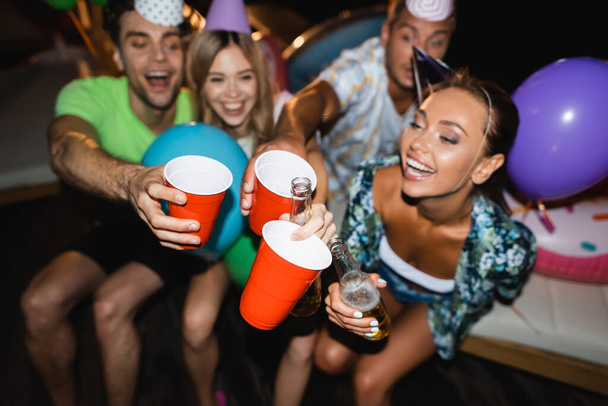 Selective focus of young friends in party caps clinking with beer and disposable cups at night  - Photo, Image