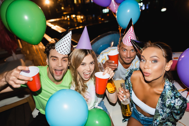 Freunde halten bei Party nachts Einwegbecher und Bierflaschen in der Nähe von Luftballons  - Foto, Bild