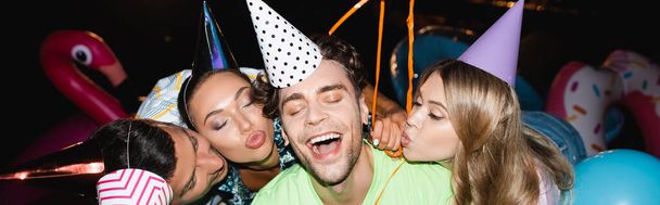 Panoramic crop of young friends kissing man in party cap near balloons at night  - Photo, Image