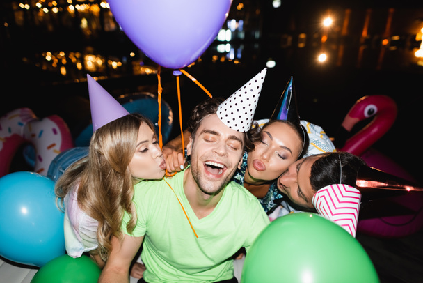 Selective focus of friends in party caps kissing man with balloons outdoors at night  - Foto, imagen