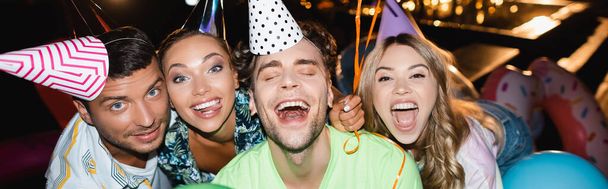 Заголовок сайта взволнованных друзей в шапочках для вечеринок, смотрящих на камеру возле бассейна ночью  - Фото, изображение