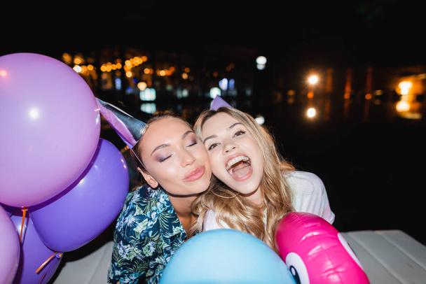 Foco seletivo de mulheres jovens em bonés de festa perto de balões à noite  - Foto, Imagem