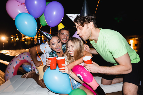Selective focus of women clinking with disposable cups near balloons during party near swimming pool at night  - Zdjęcie, obraz