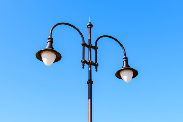 Street lamp with two bulbs against a blue sky on a sunny day. Vintage style double lamp post outdoors. Modern energy-saving technologies for lighting streets, parks and roads. Low angle view. - 写真・画像