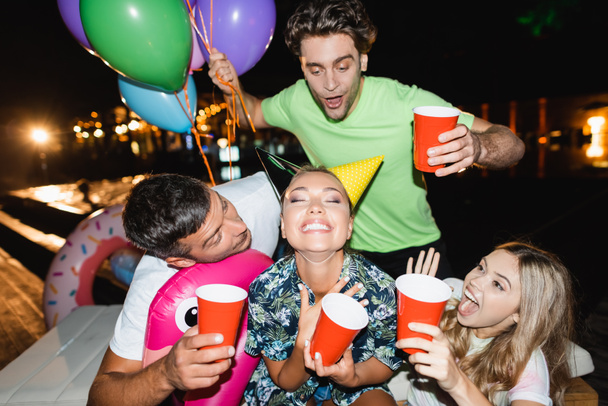 Selective focus of excited friends holding disposable cups and balloons near woman in party caps at night  - 写真・画像