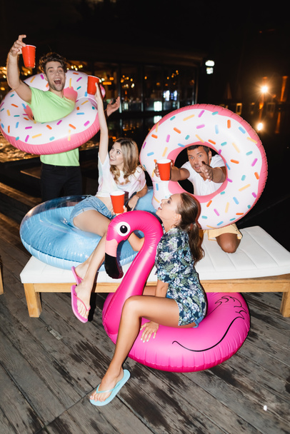 Молодые друзья с плавательными кольцами держат одноразовые чашки во время вечеринки ночью  - Фото, изображение