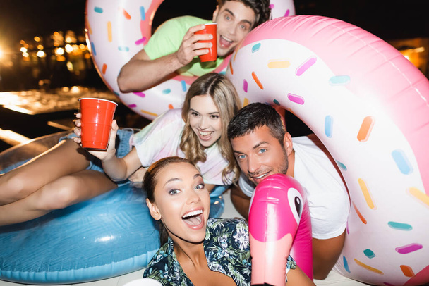 Selective focus of excited friends with swim rings holding disposable cups during party at night  - 写真・画像
